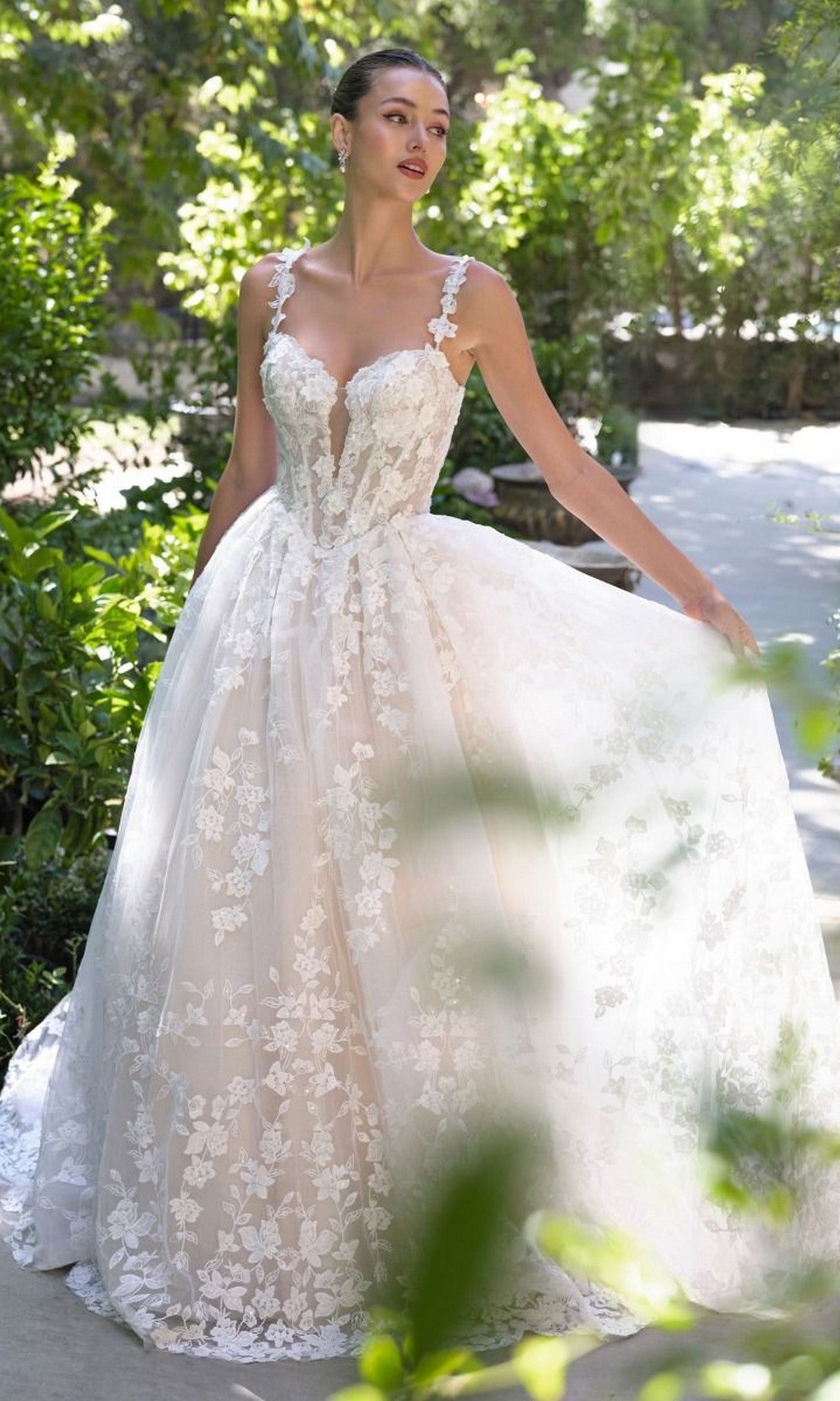 A woman poses outdoors in the Andrea and Leo WL058 long wedding dress, an a-line gown with white floral lace, thin straps, and a full skirt, surrounded by greenery and sunlight.